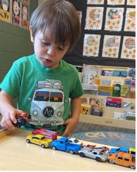 Young child in green shirt lines up toy cars on a table, focused expression.