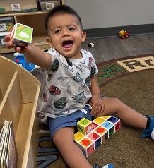 Smiling child with blocks, indoors. He's holding a block and sitting near a wooden shelf and rug.