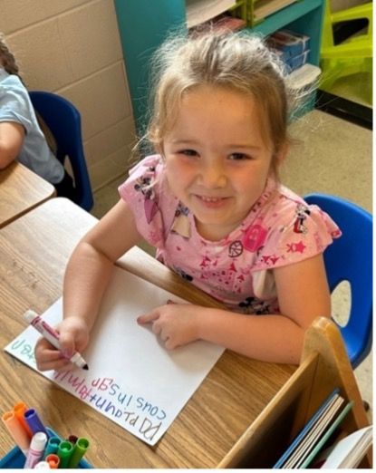 Girl at desk writing on paper, smiling. Pink shirt. Colorful markers nearby.