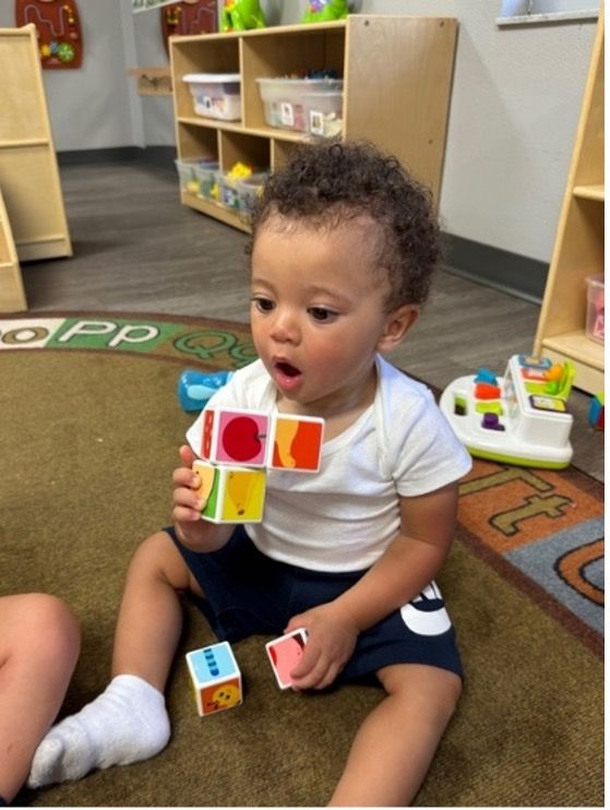 Young child sits with blocks, looking amazed in a classroom.