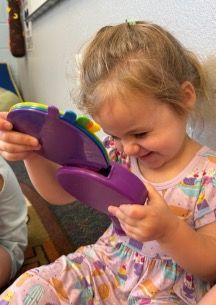 Young girl in a patterned dress smiles as she plays with a purple toy indoors.