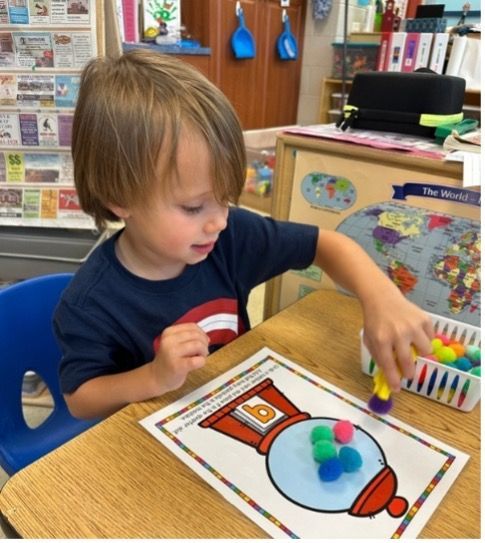 Young boy places colorful pom-poms on a worksheet of a gumball machine at a desk in a classroom.