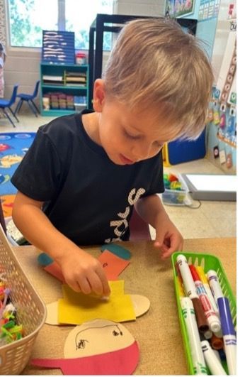 A young boy in a black shirt colors paper cutouts at a table in a classroom setting.