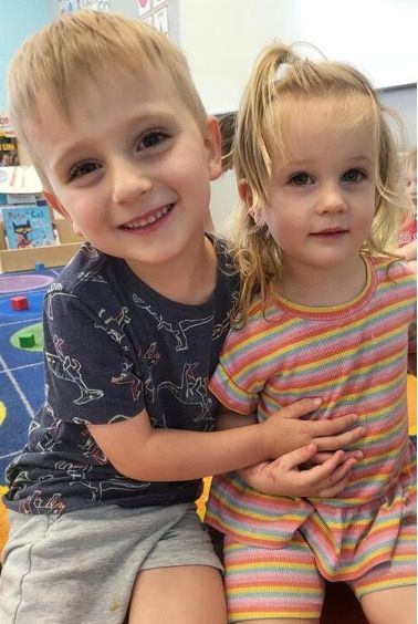 Boy with blue shirt hugs girl in striped top; both smile indoors.