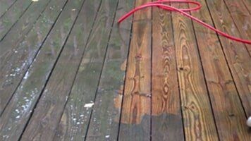 Wooden deck being power washed; half cleaned, half dirty with a red hose visible.