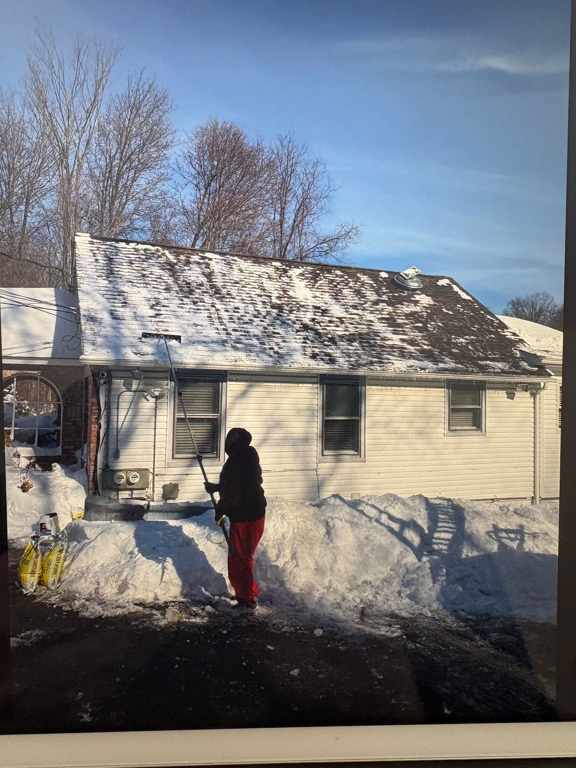 Person shoveling snow in front of a snow-covered house on a sunny day.