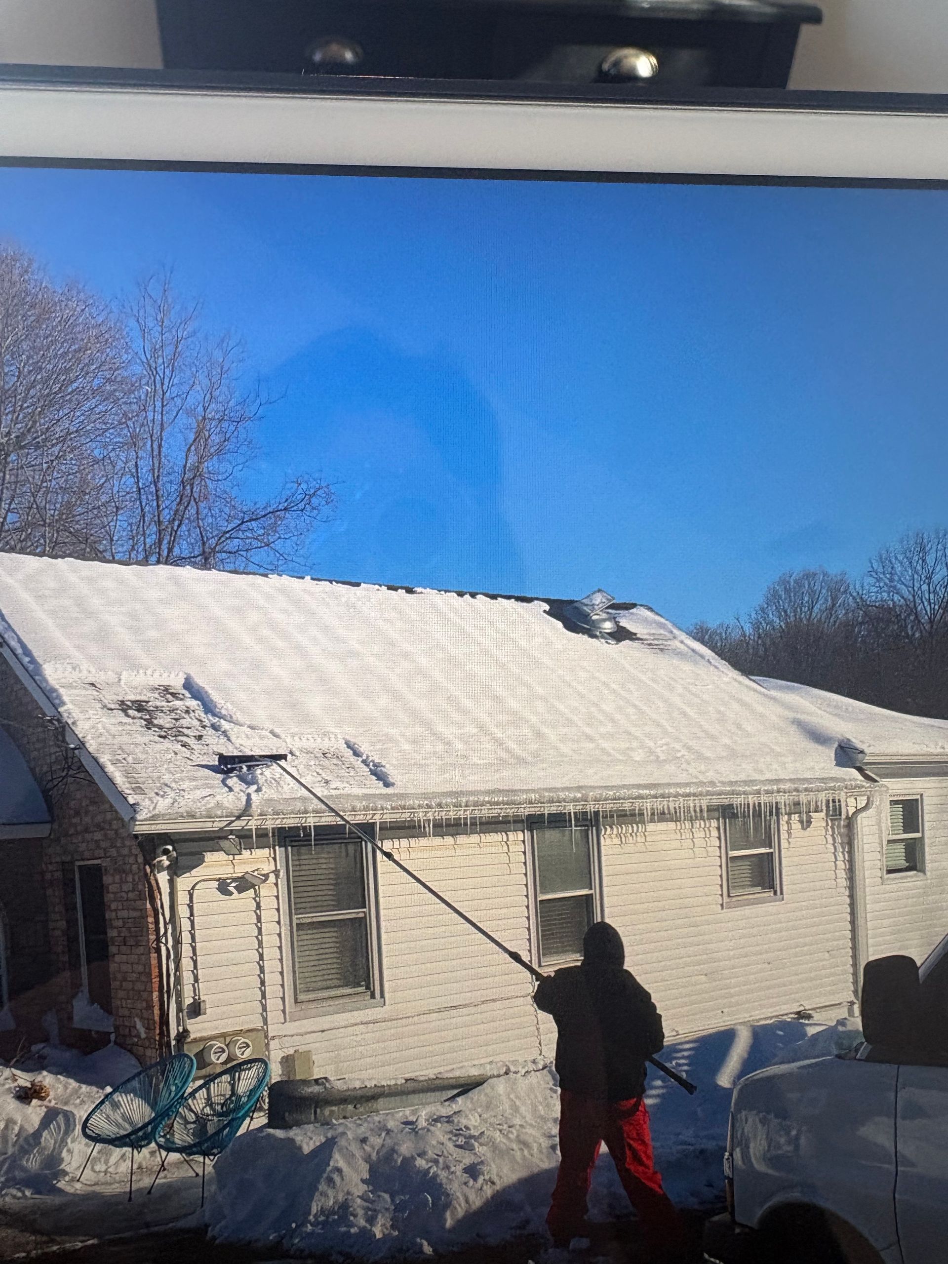 Person removes snow from a roof with a long-handled tool on a sunny, snowy day. White house, blue sky.