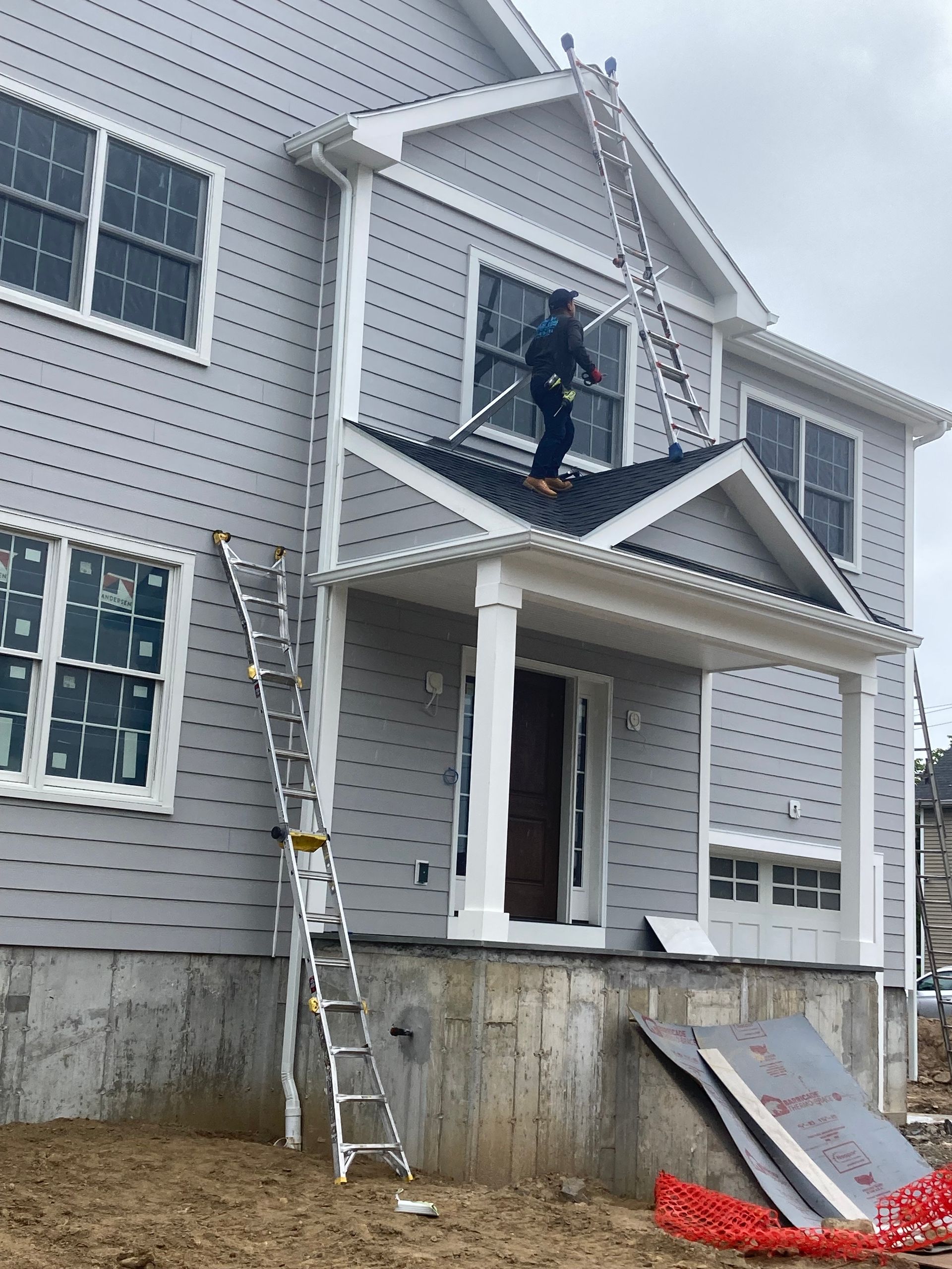 Person on a ladder working on the exterior of a two-story gray house under construction; cloudy day.