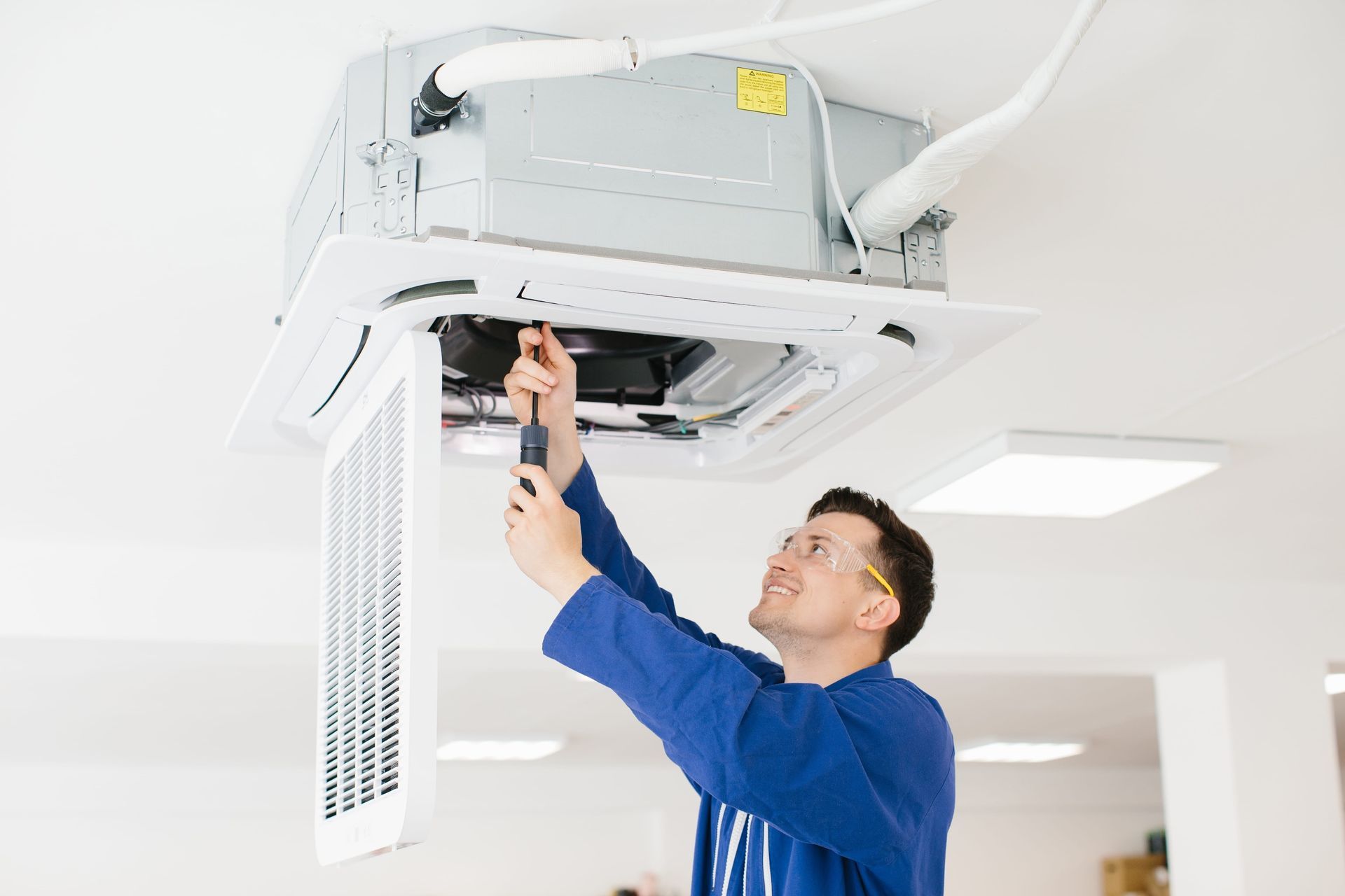 Technician in blue shirt servicing a ceiling-mounted HVAC unit in a white room