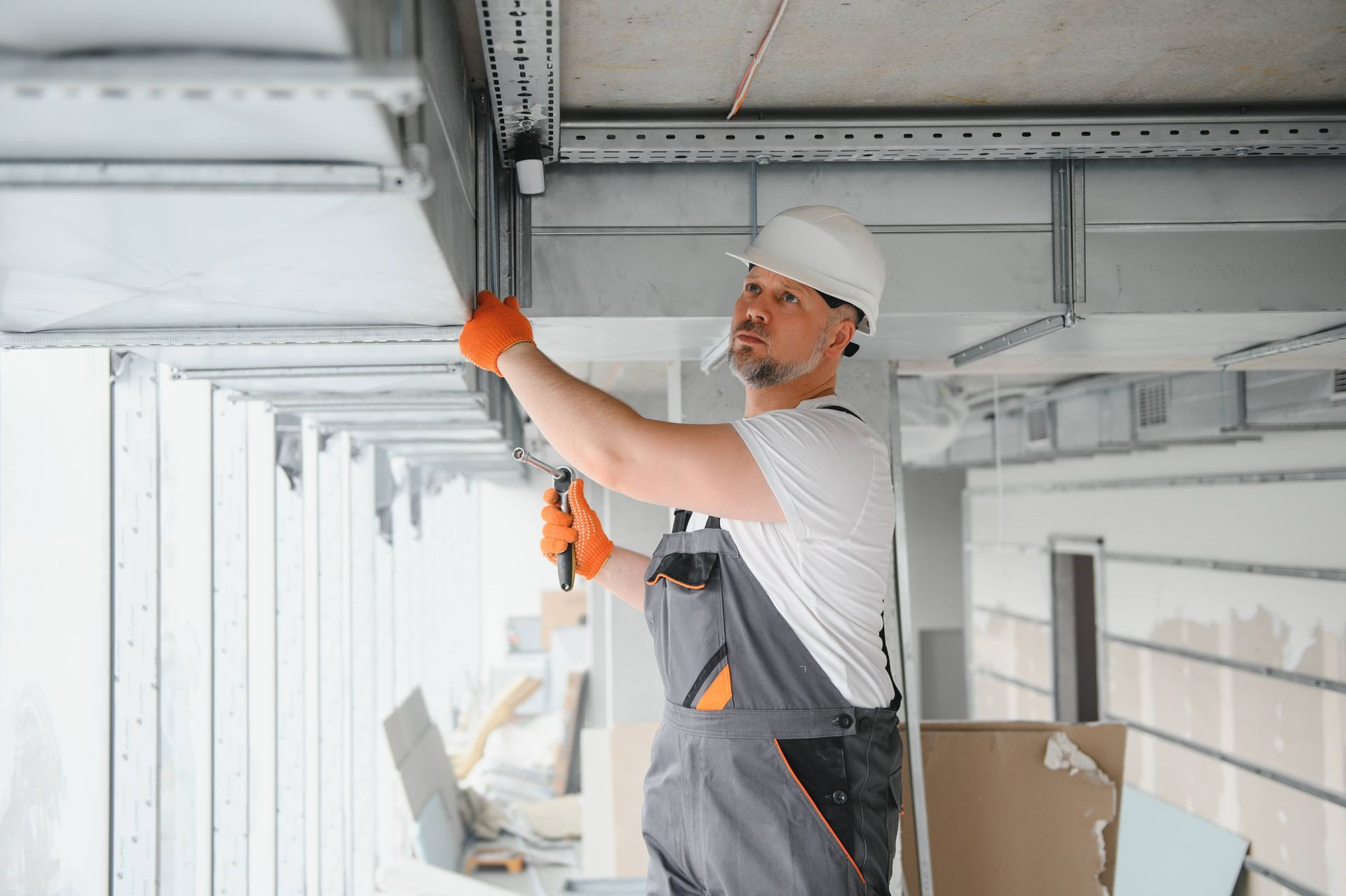 Worker in hard hat and orange gloves adjusting overhead metal framing in a bright construction site.