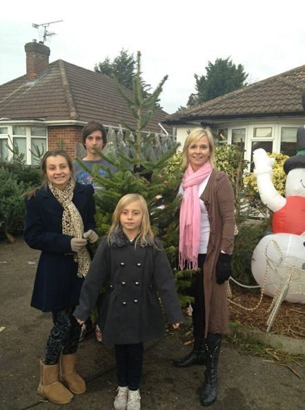 Family posing with Christmas tree outside a house. Woman in brown coat, pink scarf, and boots. Children in winter coats.