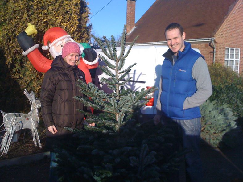 Two people pose next to a Christmas tree outdoors; inflatable Santa and reindeer decoration in background.