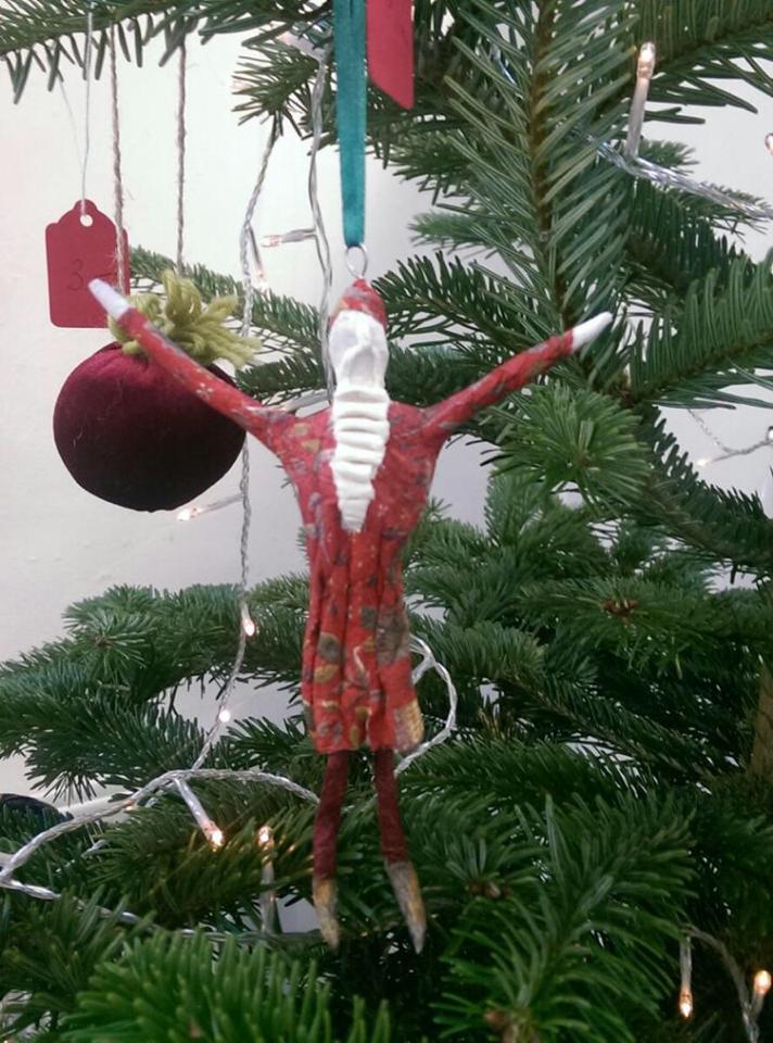 Christmas tree ornament of Santa with arms outstretched, red and white, hanging from a green tree.