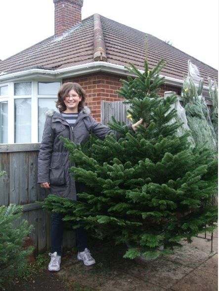 Woman beside a fresh-cut Christmas tree, smiling. Standing in front of a house, brick exterior, and wooden fence.