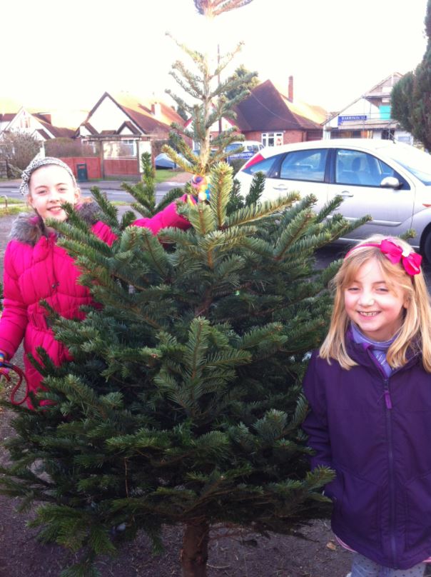 Two girls smile next to a small Christmas tree outdoors. One wears a pink jacket, the other purple.