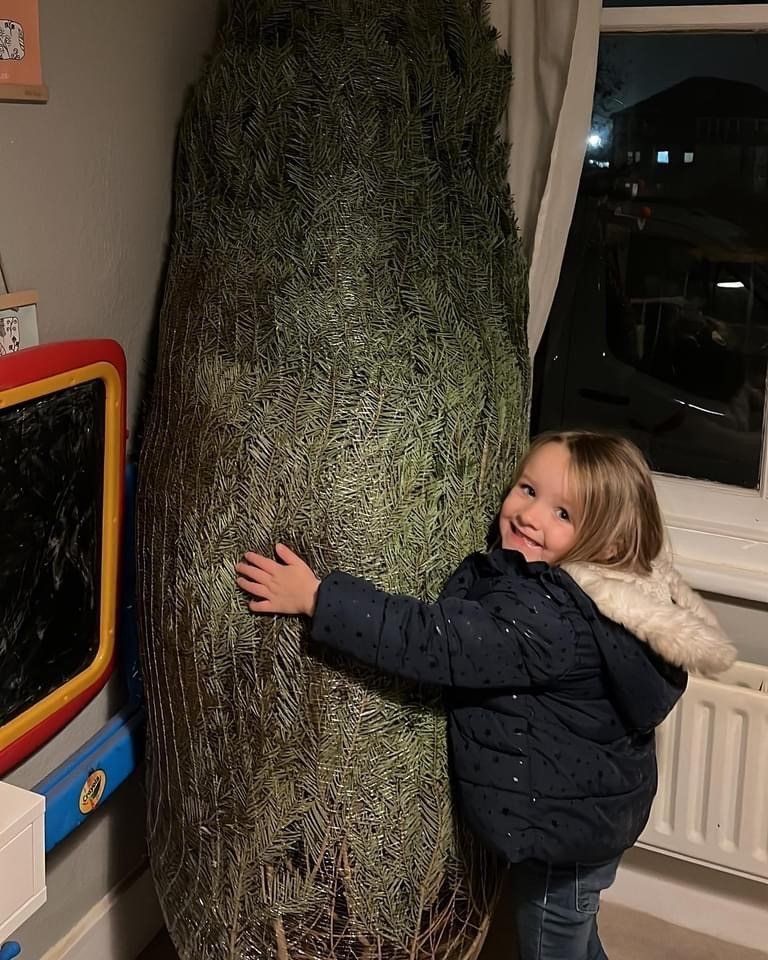Young child hugging a tall, wrapped Christmas tree indoors, smiling.