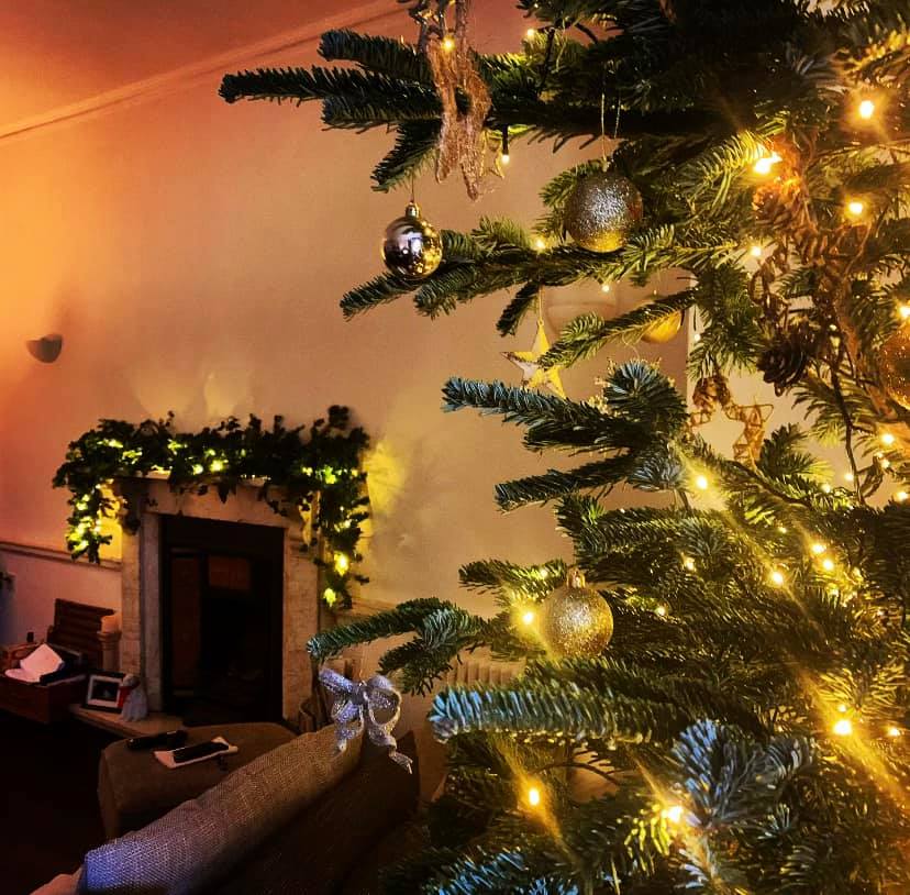 Christmas tree decorated with gold ornaments and lights, beside a fireplace with garland.