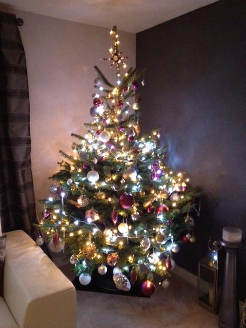 Decorated Christmas tree with ornaments and lights in a living room; dark wall and sofa visible.