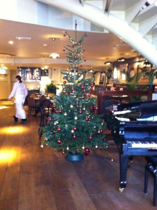 Christmas tree in restaurant, decorated with red and gold ornaments. A waiter walks by, a piano is nearby.