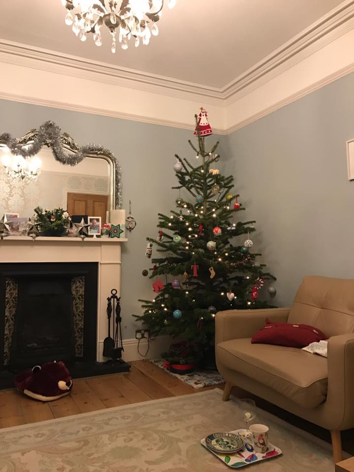 Christmas tree in a cozy room, decorated with ornaments. A fireplace and a chair are also in view.