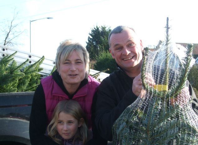 Family with a Christmas tree wrapped in netting. They are smiling, outdoors, possibly at a tree lot.