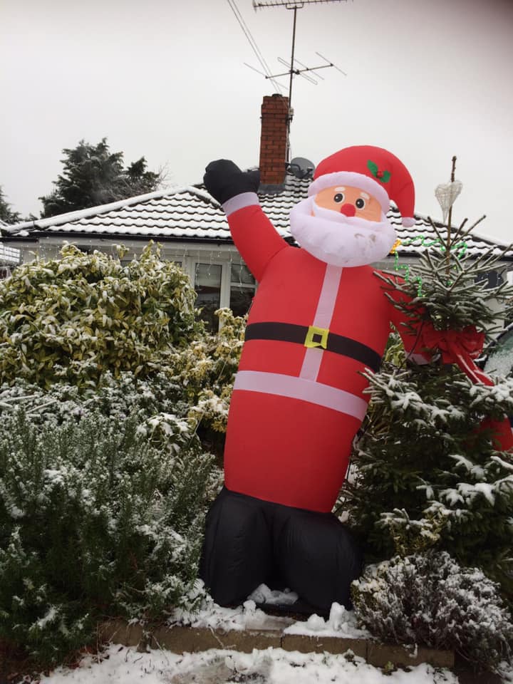 Inflatable Santa Claus waving in front of a snow-covered house and bushes.
