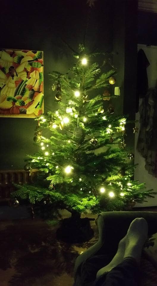 Lit Christmas tree in a dark room with glowing lights, a painting on the wall, and feet in the foreground.