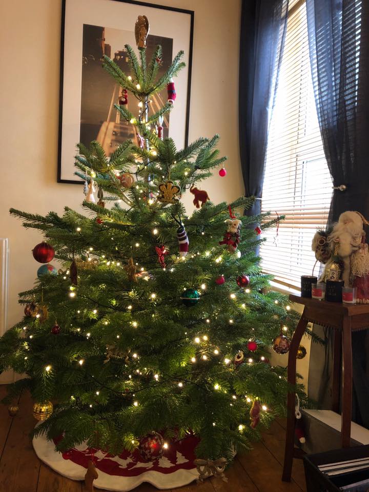 Christmas tree decorated with lights and ornaments, in front of a window and artwork.