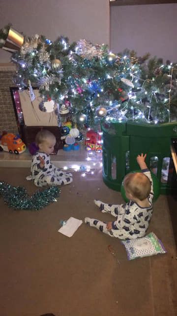 Two toddlers in pajamas play near a decorated Christmas tree with fallen tinsel and ornaments.