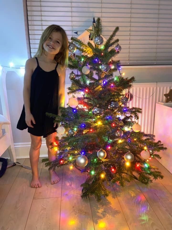 Girl in black dress smiles beside a Christmas tree decorated with ornaments and colorful lights.