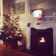 Christmas tree decorated with red ornaments next to a lit fireplace, with a Santa figurine on the mantel.