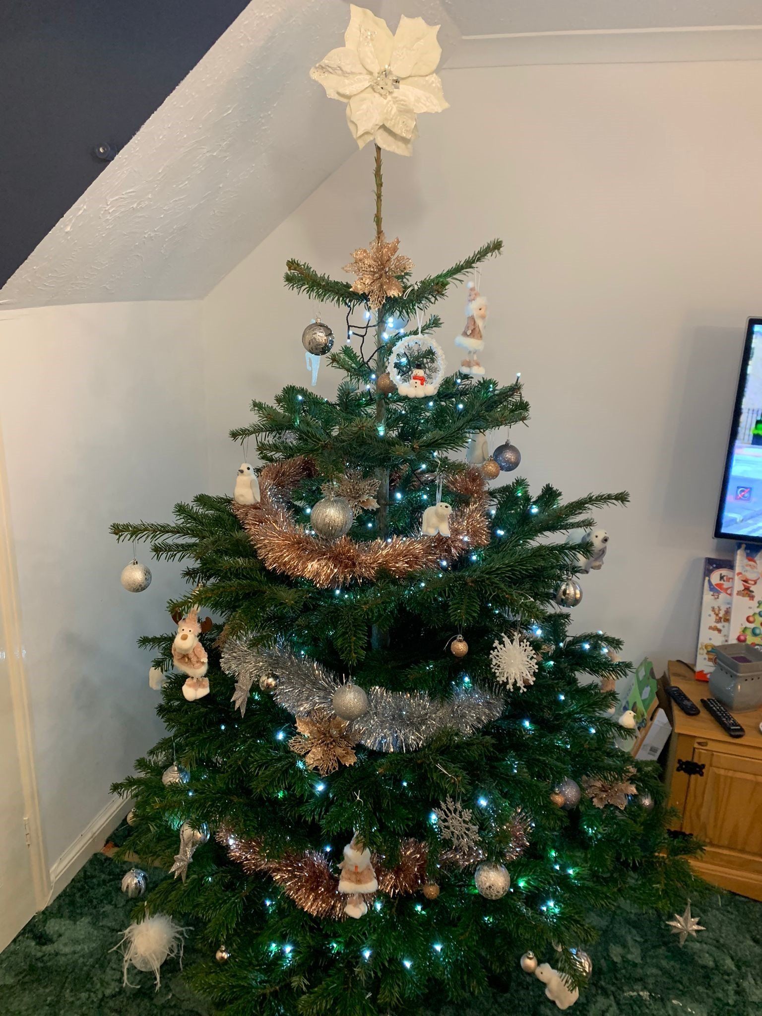 Christmas tree decorated with ornaments and garland, topped with a white flower. Lit with small green lights.