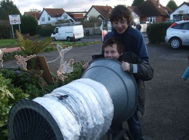 Two boys smile, positioned behind a large, makeshift cannon made of metal containers, outdoors.
