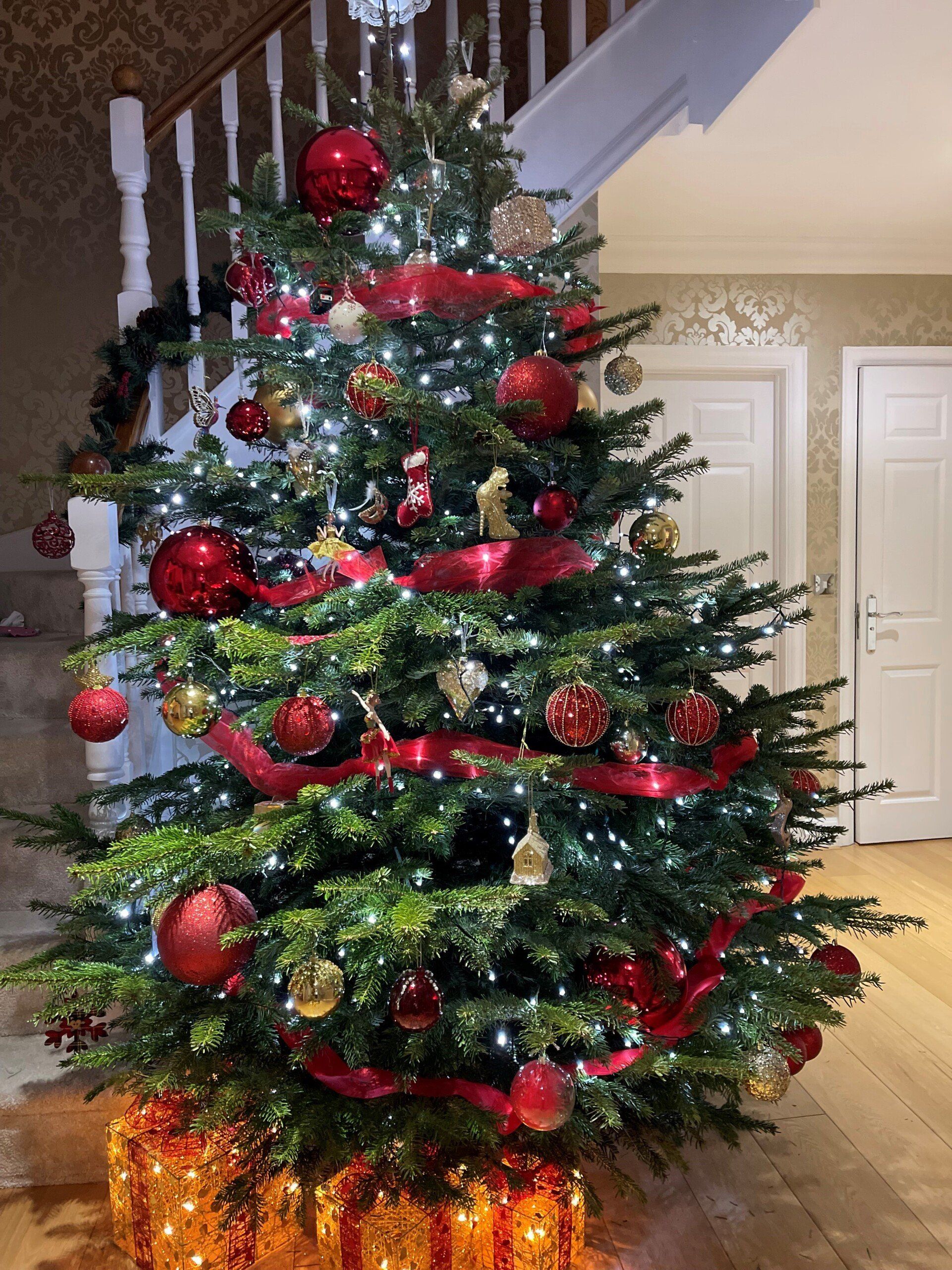 Christmas tree decorated with red ornaments, lights, and ribbon, next to a staircase.