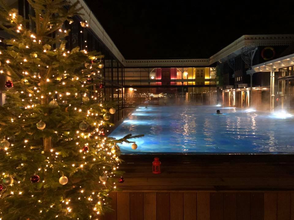 Christmas tree with lights next to an outdoor pool at night; steam rising, colorful building in background.