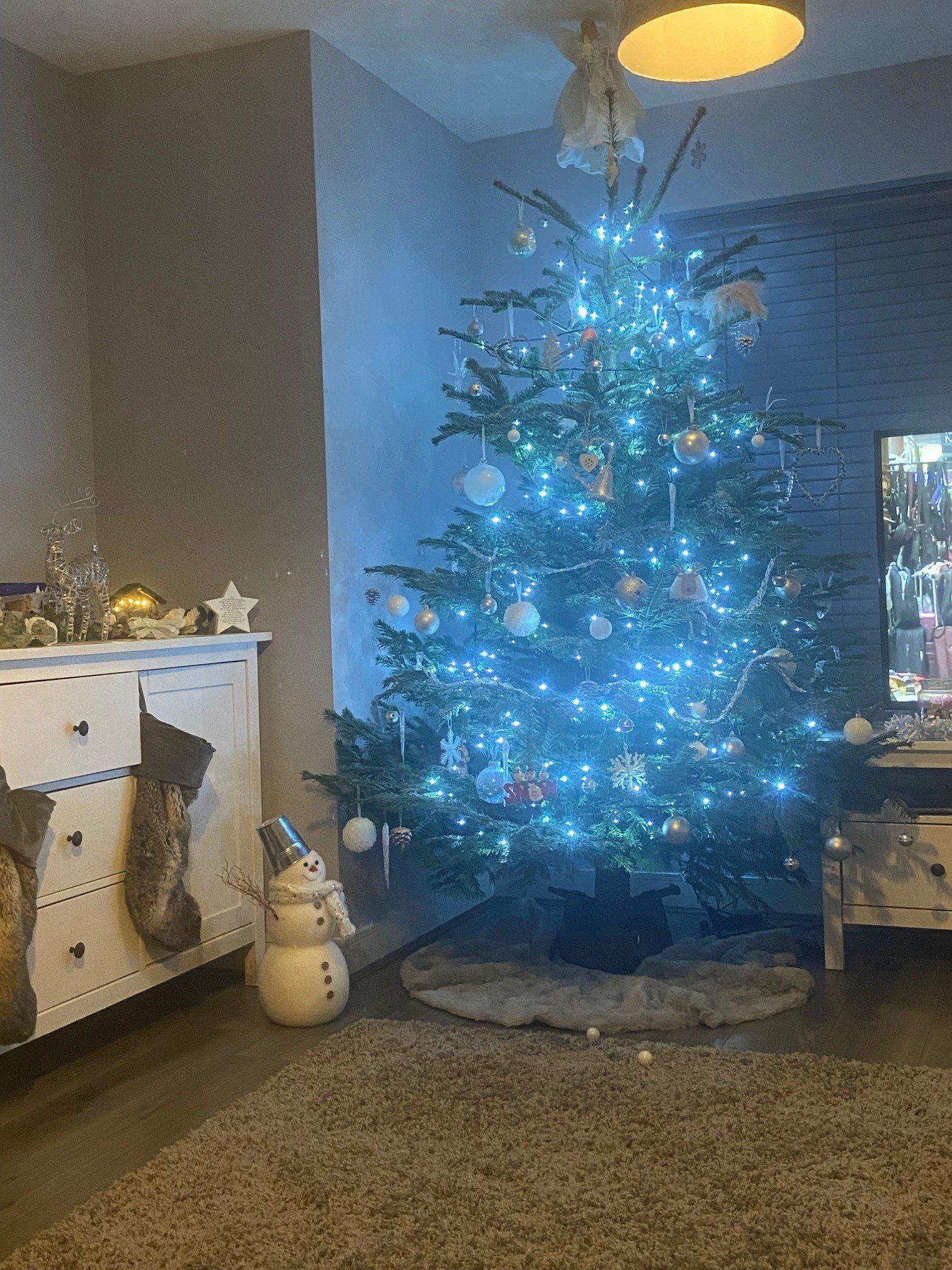 Christmas tree lit with blue lights and ornaments in a living room, next to a white dresser.