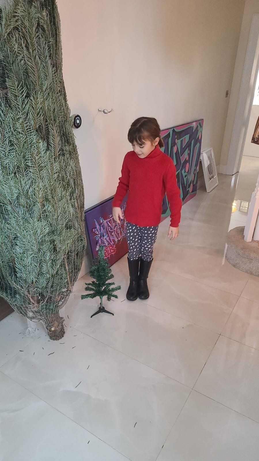 Child standing near a small artificial tree, with a large, bundled Christmas tree in the background.