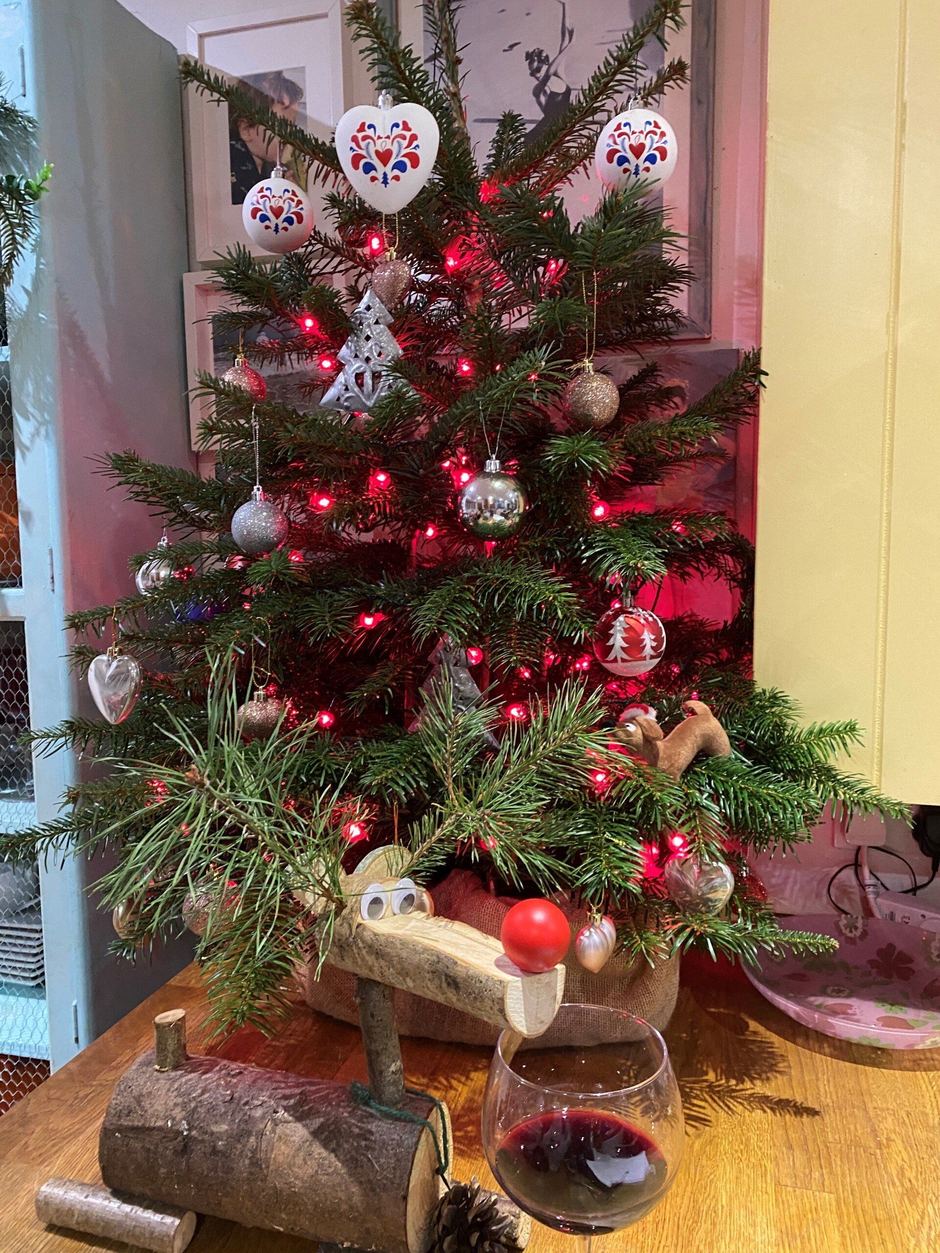 A decorated Christmas tree with red lights and ornaments sits on a table next to a glass of red wine.