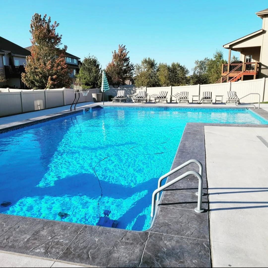 Outdoor swimming pool with blue water, concrete deck, and seating. Fence and houses in the background.