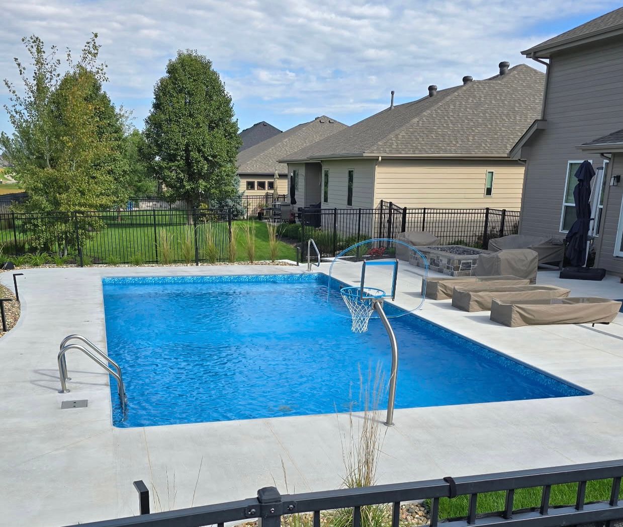 Rectangular pool with blue water and ladders, surrounded by concrete patio and houses in the background.