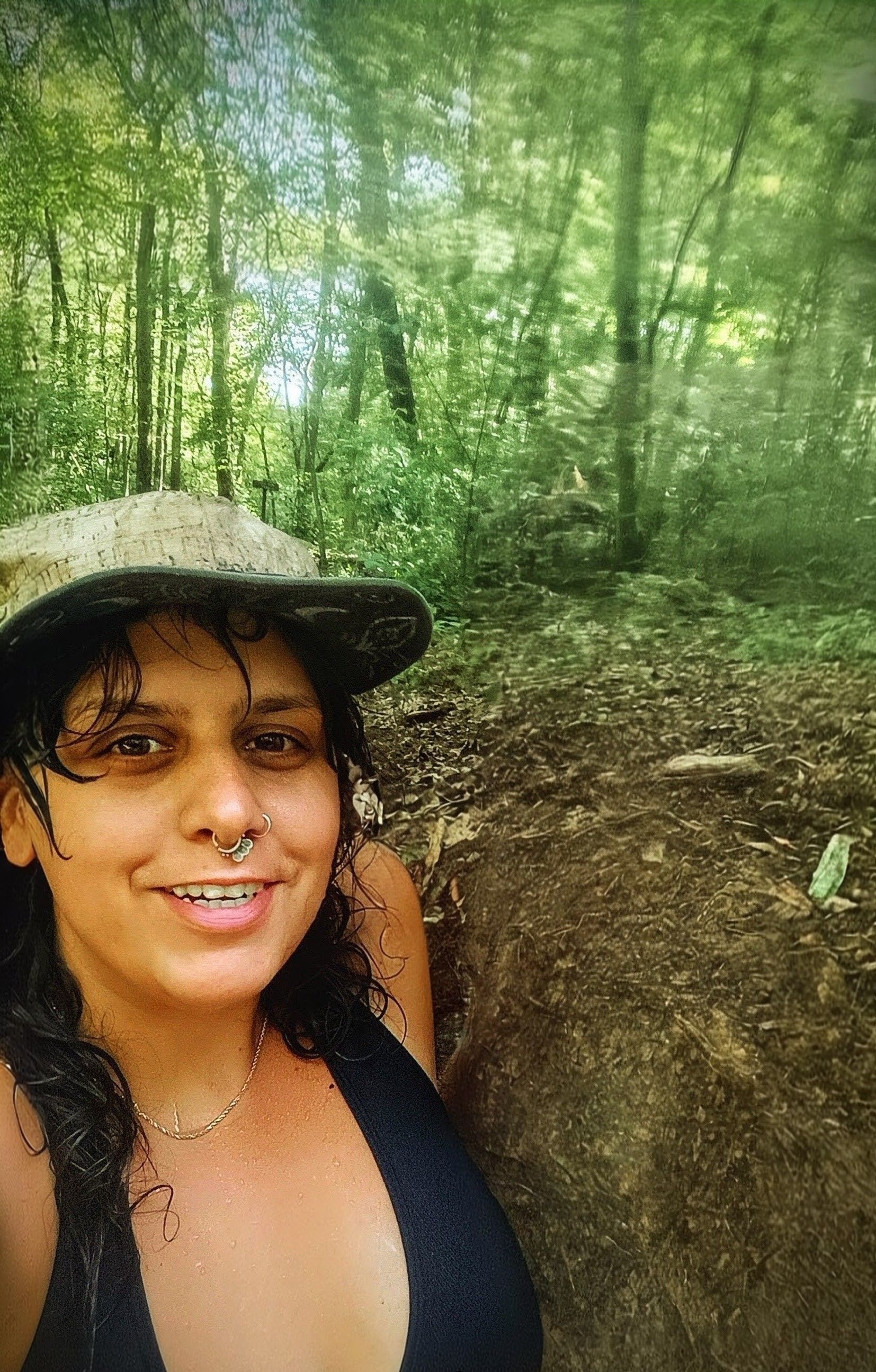 Woman in hat and black top smiles in a forest setting.