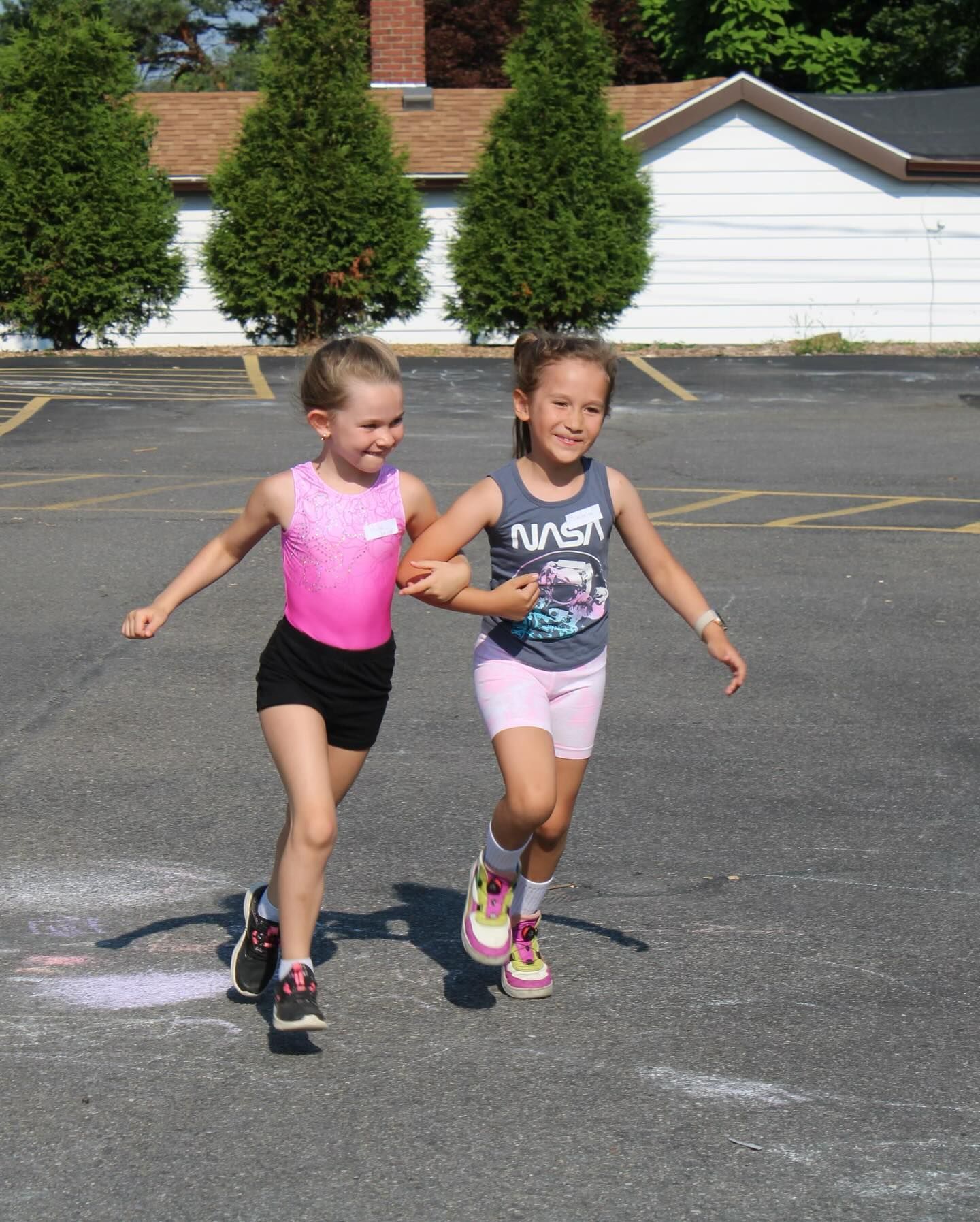 Two young girls, arm-in-arm, run across an asphalt parking lot, smiling. One wears pink, the other blue.