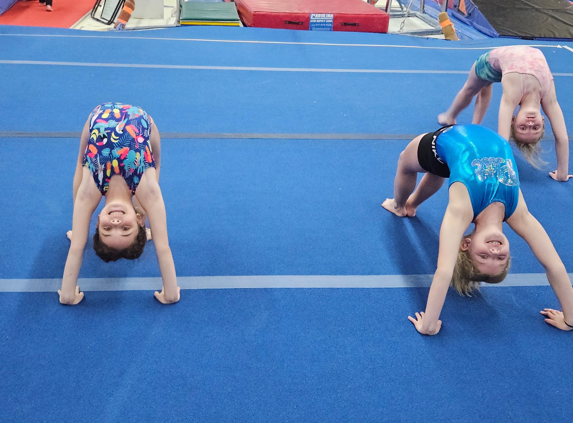 Three young gymnasts in leotards do backbends on a blue mat in a gym, smiling at the camera.