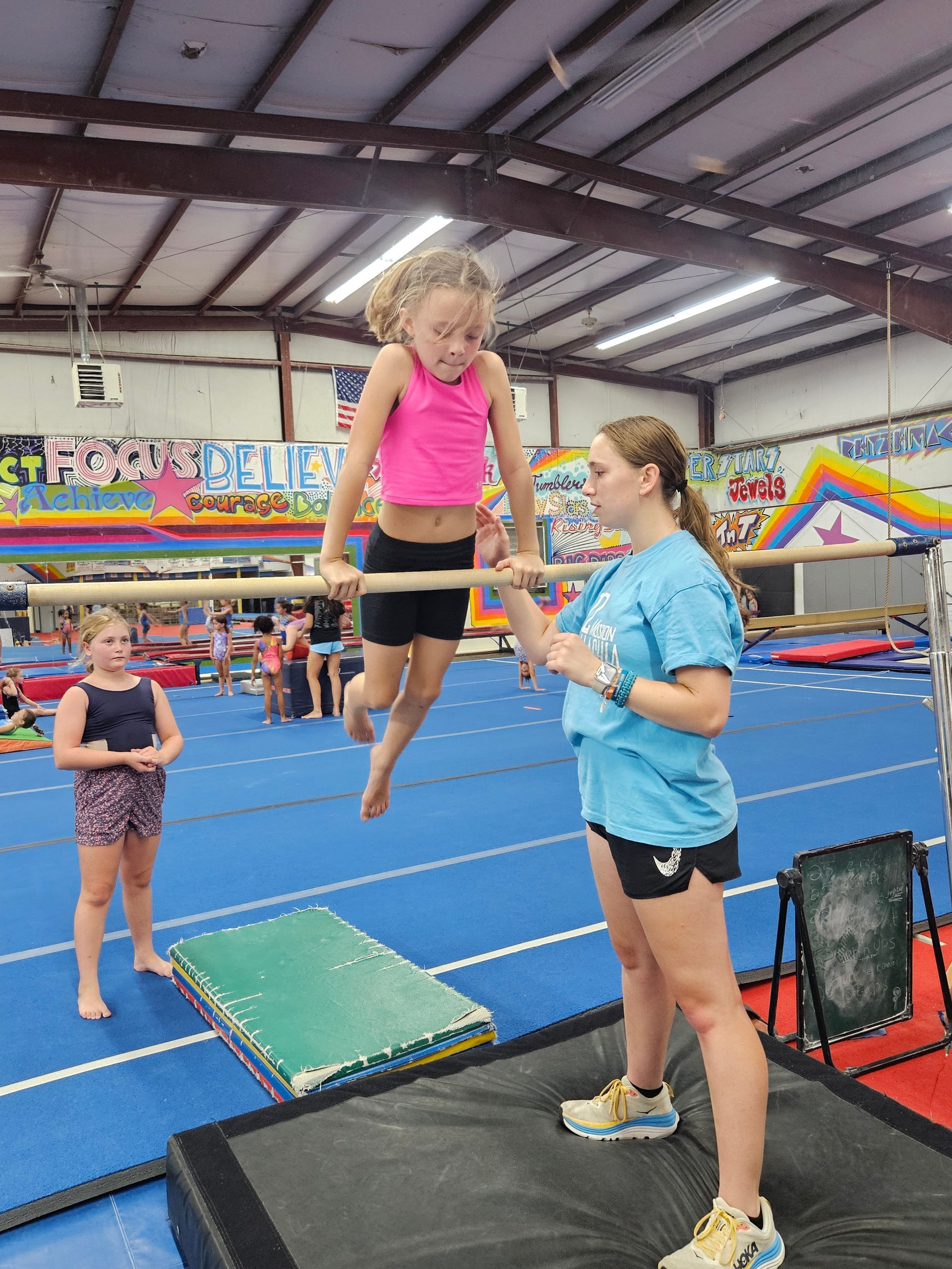 Girl on gymnastics bar, assisted by instructor. Gym setting, blue mats, other children present.