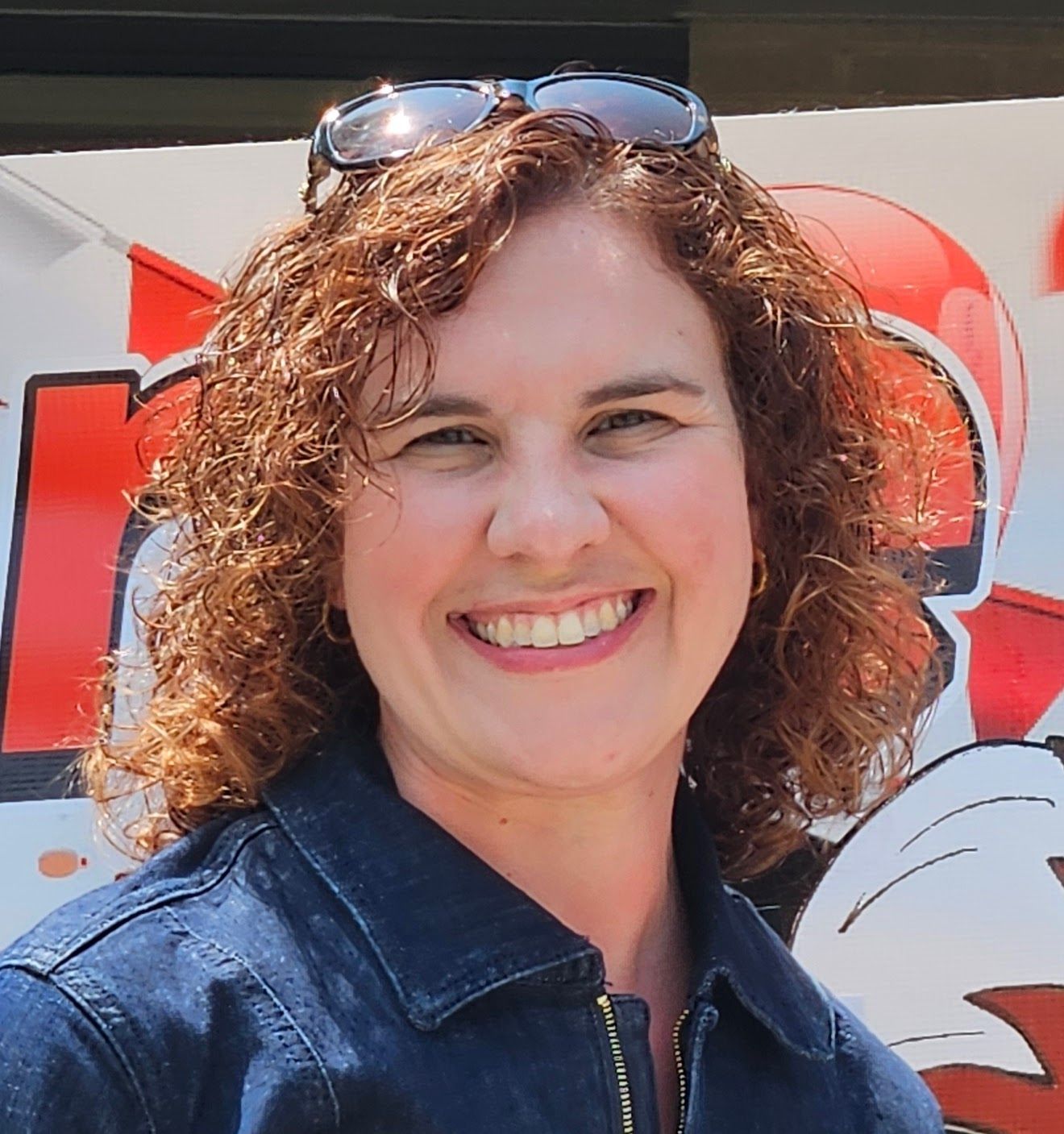 Woman with curly brown hair smiles, wearing sunglasses, and a denim jacket.