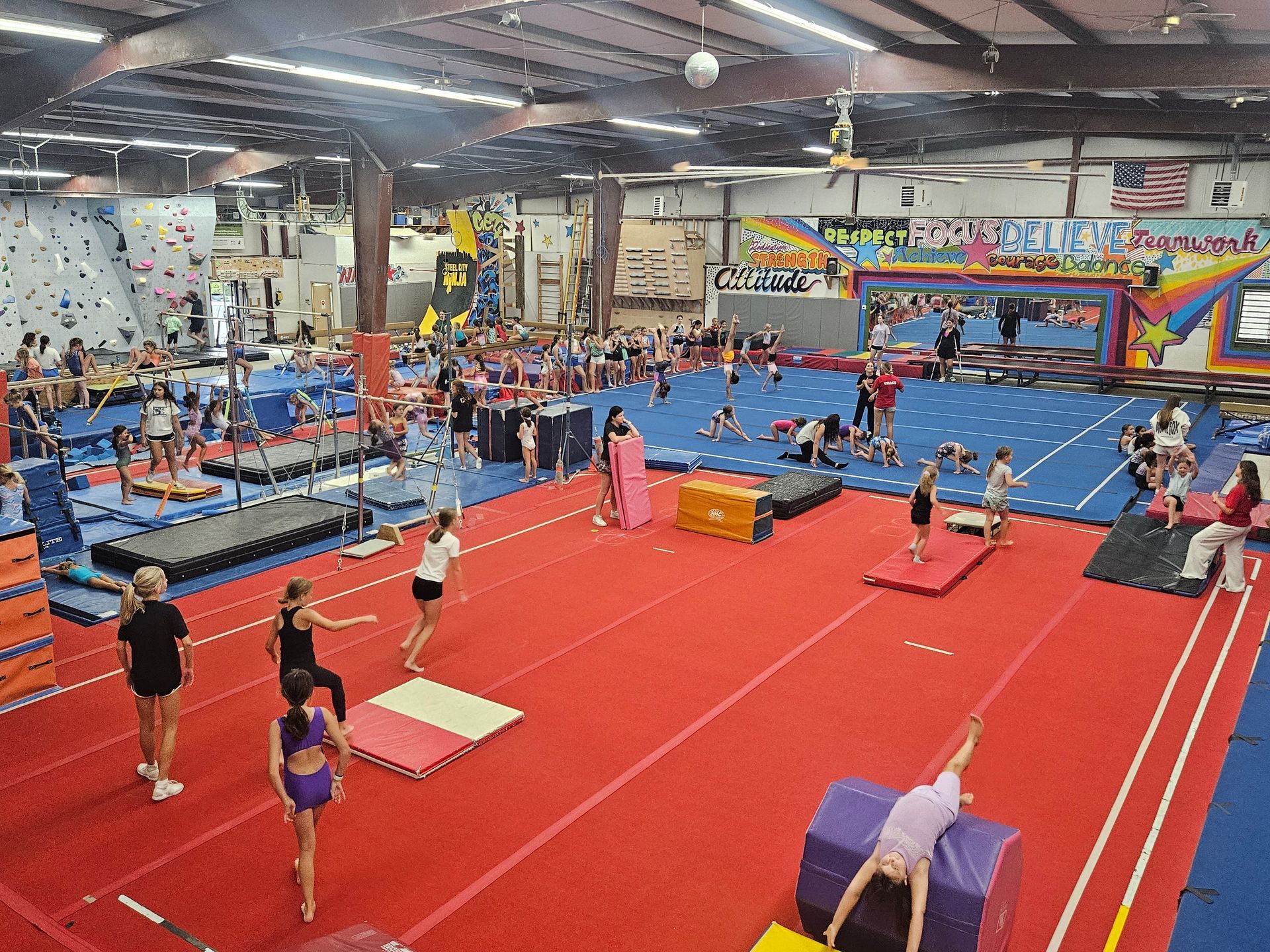 Gymnastics gym with kids practicing various skills on mats, bars, and a climbing wall.