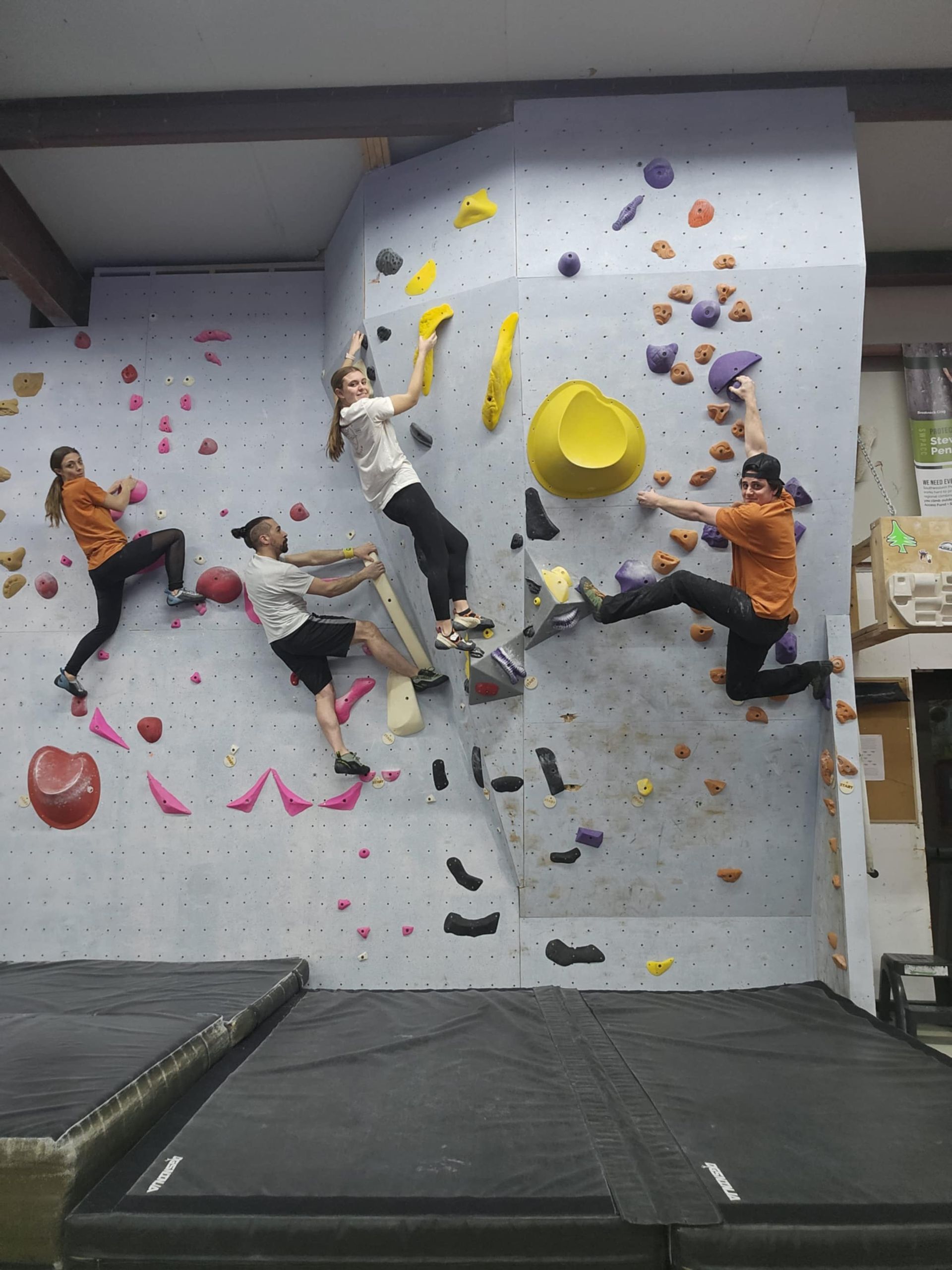 Four people bouldering on an indoor climbing wall, reaching for holds.