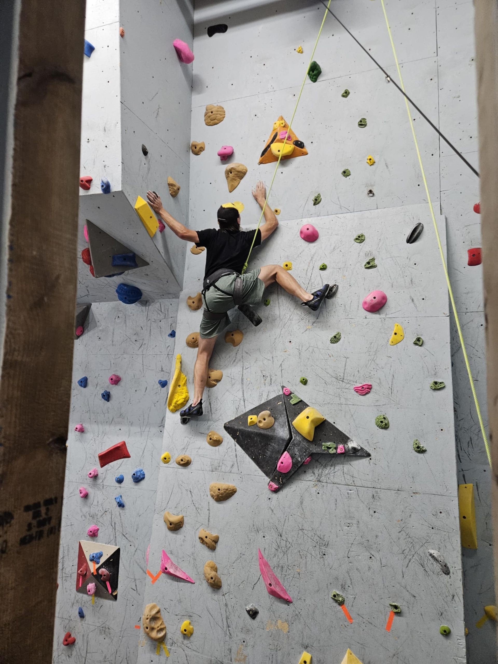 A person climbs a rock wall indoors. They wear a harness, shorts, and black shirt, reaching for a yellow hold.