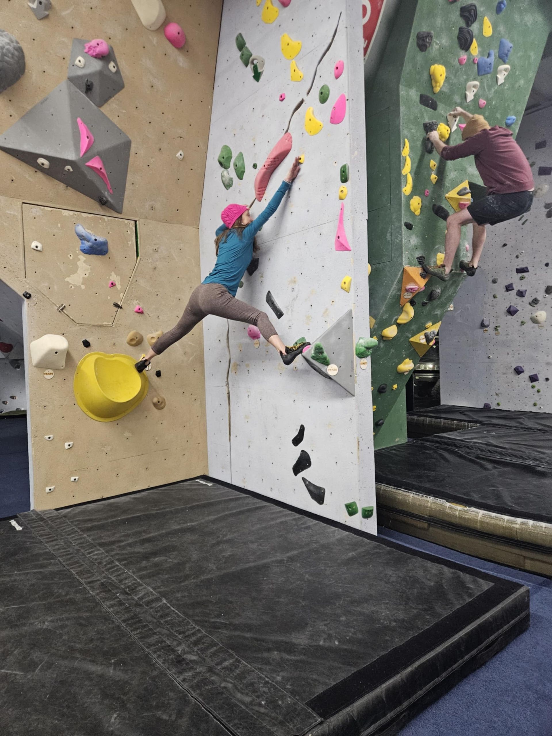 Woman rock climbing indoors, legs split, reaching for hold. Another person climbs nearby.