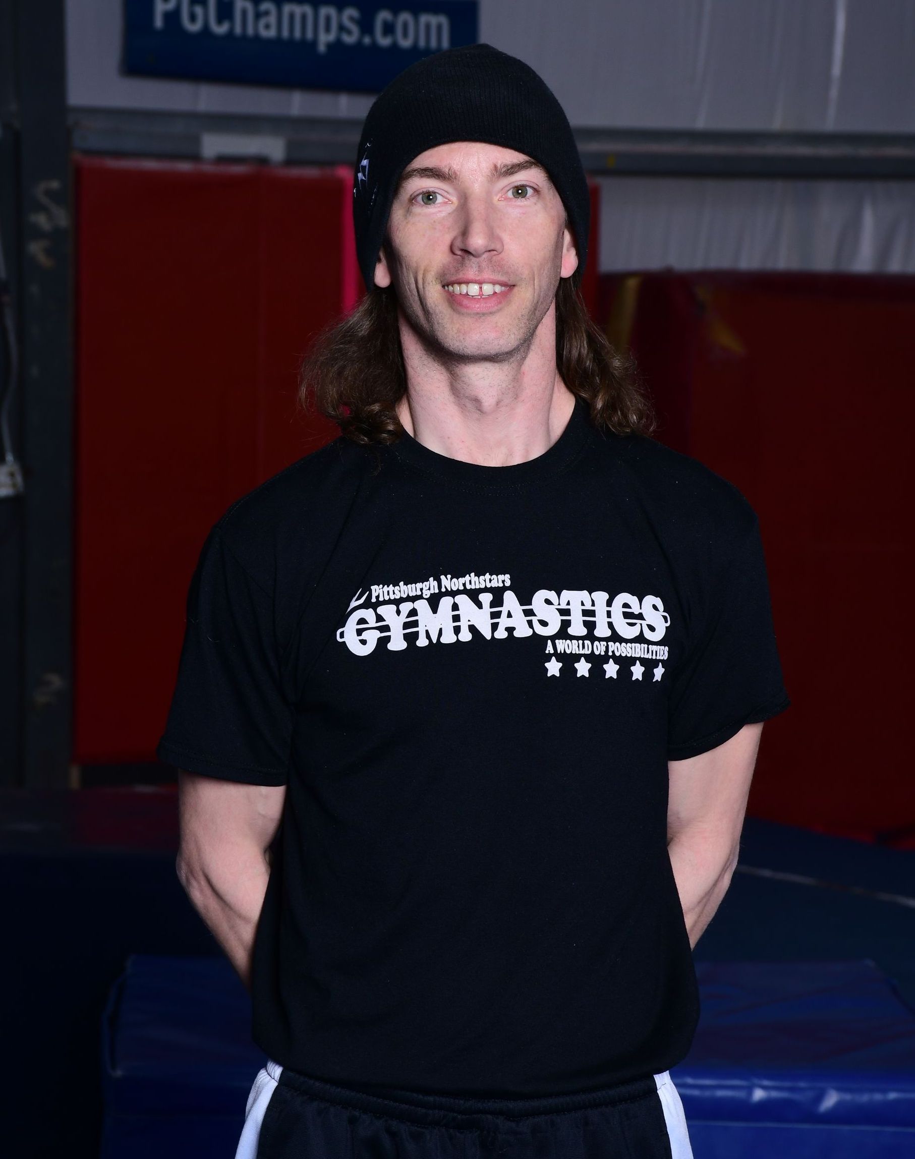 Man with long hair wearing a black hat and shirt, standing in a gym.